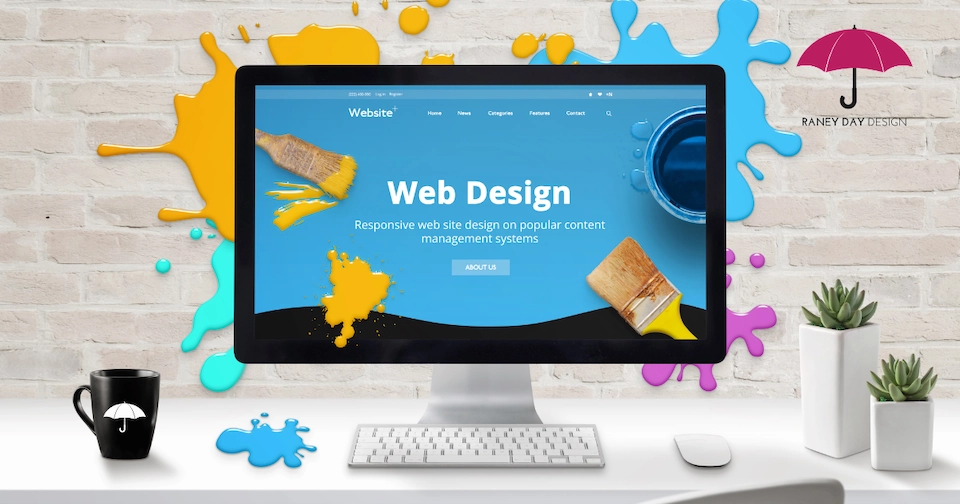 A desktop computer displays a bright blue web design homepage with the text 'Web Design – Responsive web site design on popular content management systems.' Colorful paint splashes in yellow, blue, and pink decorate the white brick wall behind the monitor.