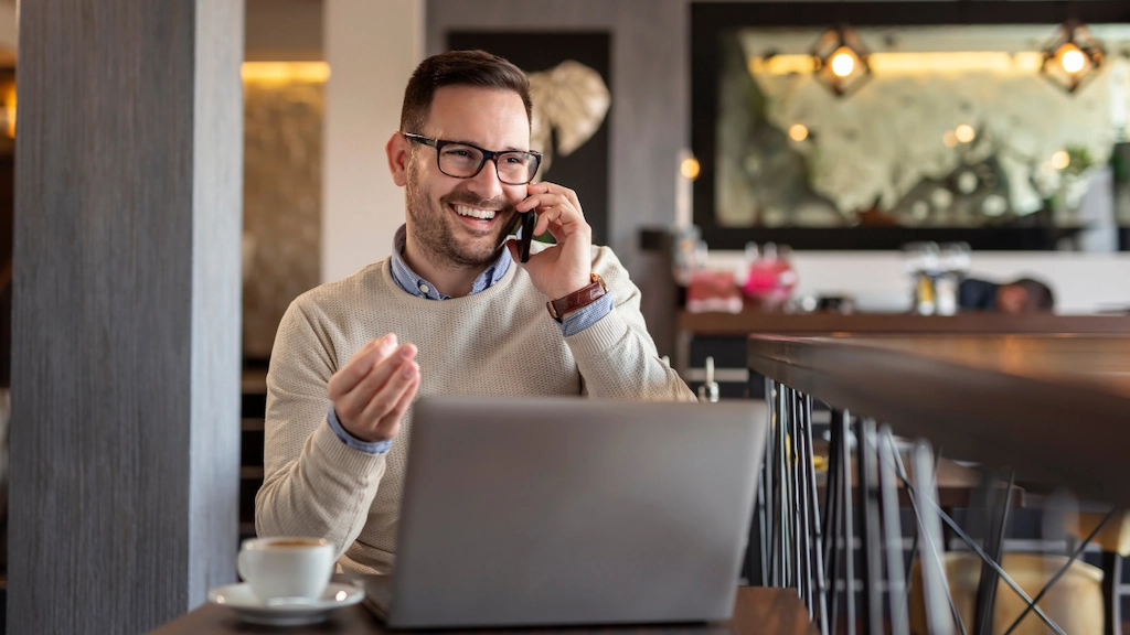a man is smiling while on the phone, sitting at a table with a cup of coffee and his laptop open